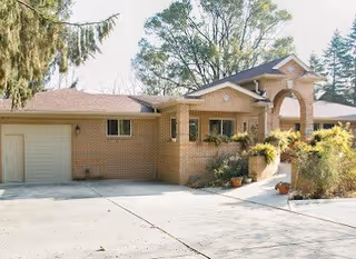Exterior view of a single-story brick building with a garage on the left side, a covered entrance with an archway, and surrounding trees and plants.