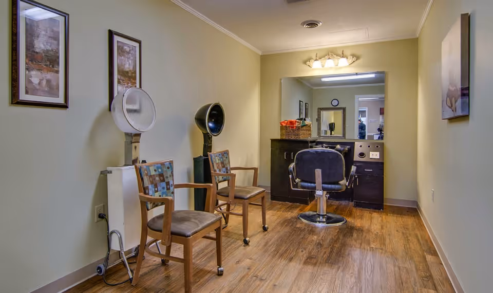 Interior view of a small salon area in a senior living facility with two wooden chairs with patterned cushions, a black salon chair in front of a large mirror, a hair dryer, and framed artwork on the walls. The floor is wooden and the walls are painted light yellow.