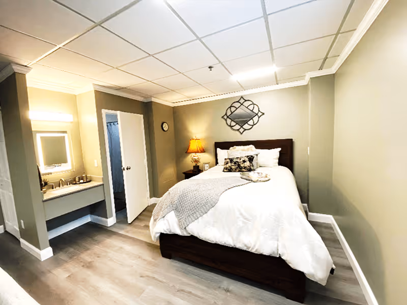 A cozy senior living bedroom with a neatly made bed featuring white and gray bedding, a decorative pillow, and a tray. The room has light green walls, a wooden nightstand with a lamp, a wall clock, and a decorative mirror above the bed. To the left, there is an open door leading to a bathroom area with a sink and mirror.