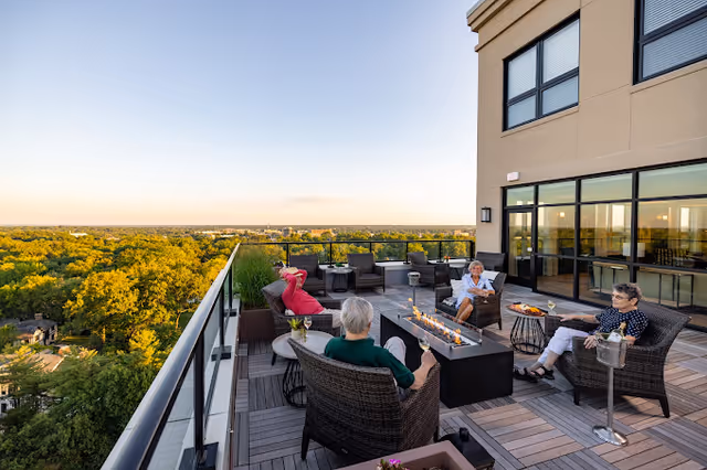 Rooftop terrace with wicker seating and a long fire pit where several residents relax, overlooking a tree-lined horizon.