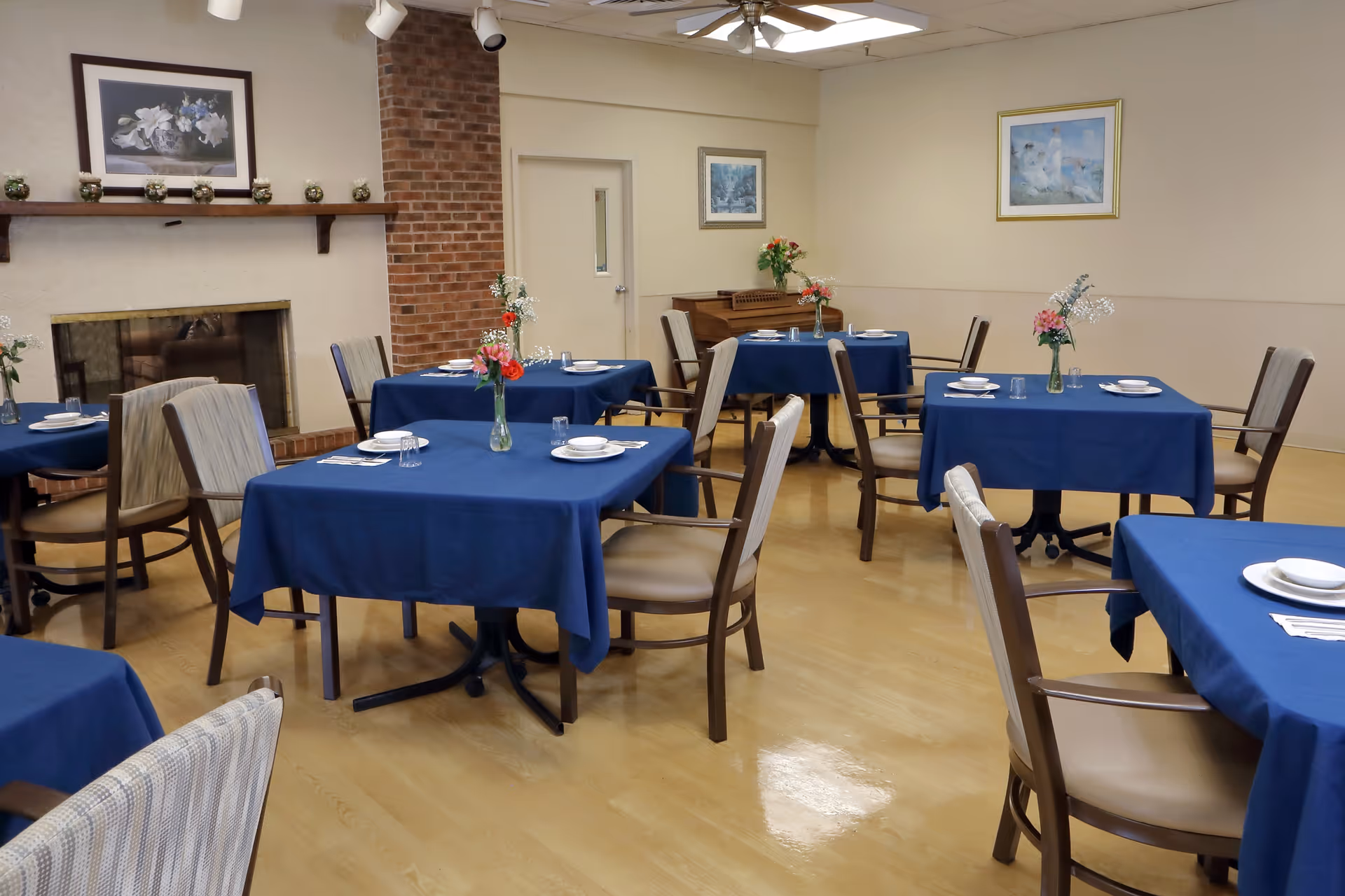 A dining room with several square tables covered with blue tablecloths, each set with plates, glasses, and silverware. Each table has a vase with flowers. The room has wooden chairs with cushioned seats, a brick fireplace, framed artwork on the walls, and a piano in the corner.