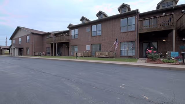 Front exterior of a two-story brick senior living building with balconies, an American flag, and a paved driveway.