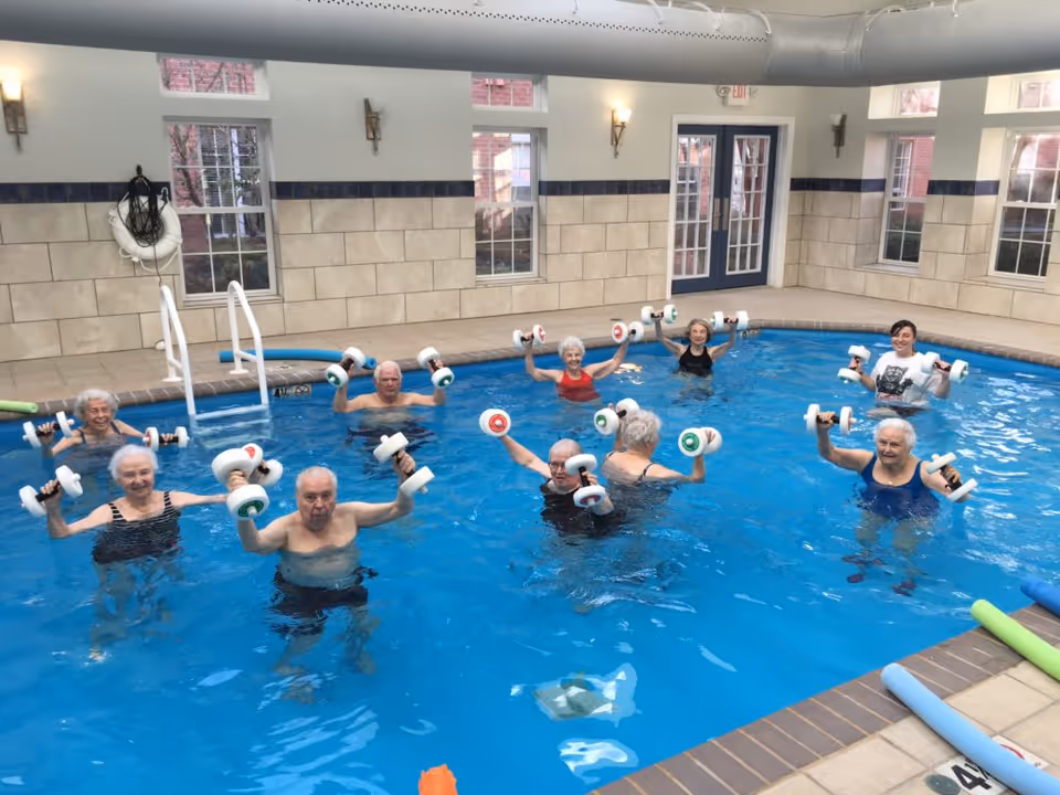 A group of elderly people participating in a water exercise class in an indoor swimming pool, each holding white foam dumbbells. The pool area has beige tiled walls, several windows, and a door leading outside.