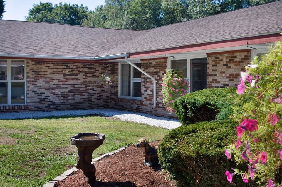 Courtyard of a single-story brick building with hanging flower baskets, trimmed shrubs, a birdbath and a small dog near the garden beds.