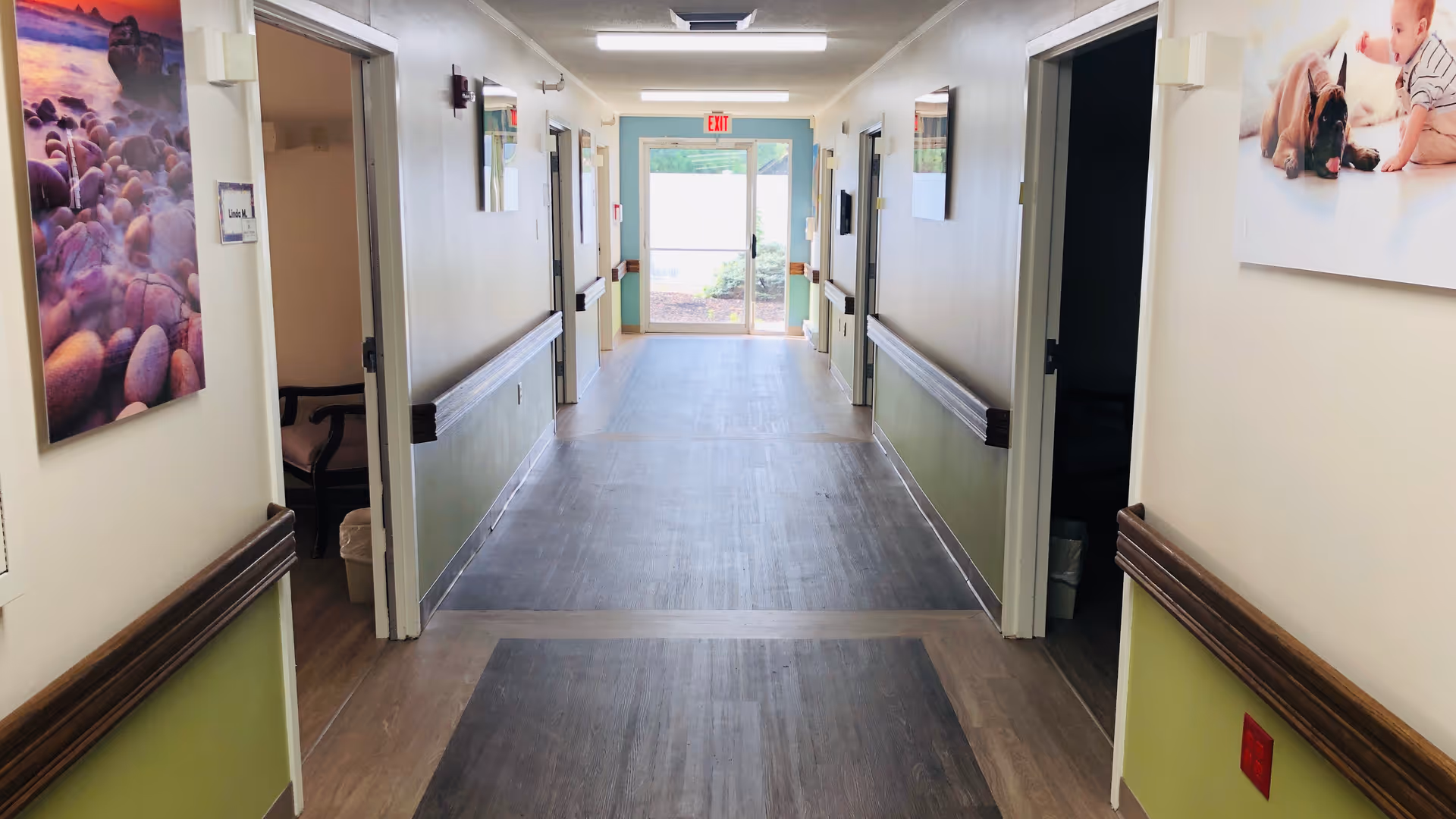 Long, well-lit nursing home hallway with wood-look flooring, handrails, open room doors, and an exit door at the far end.