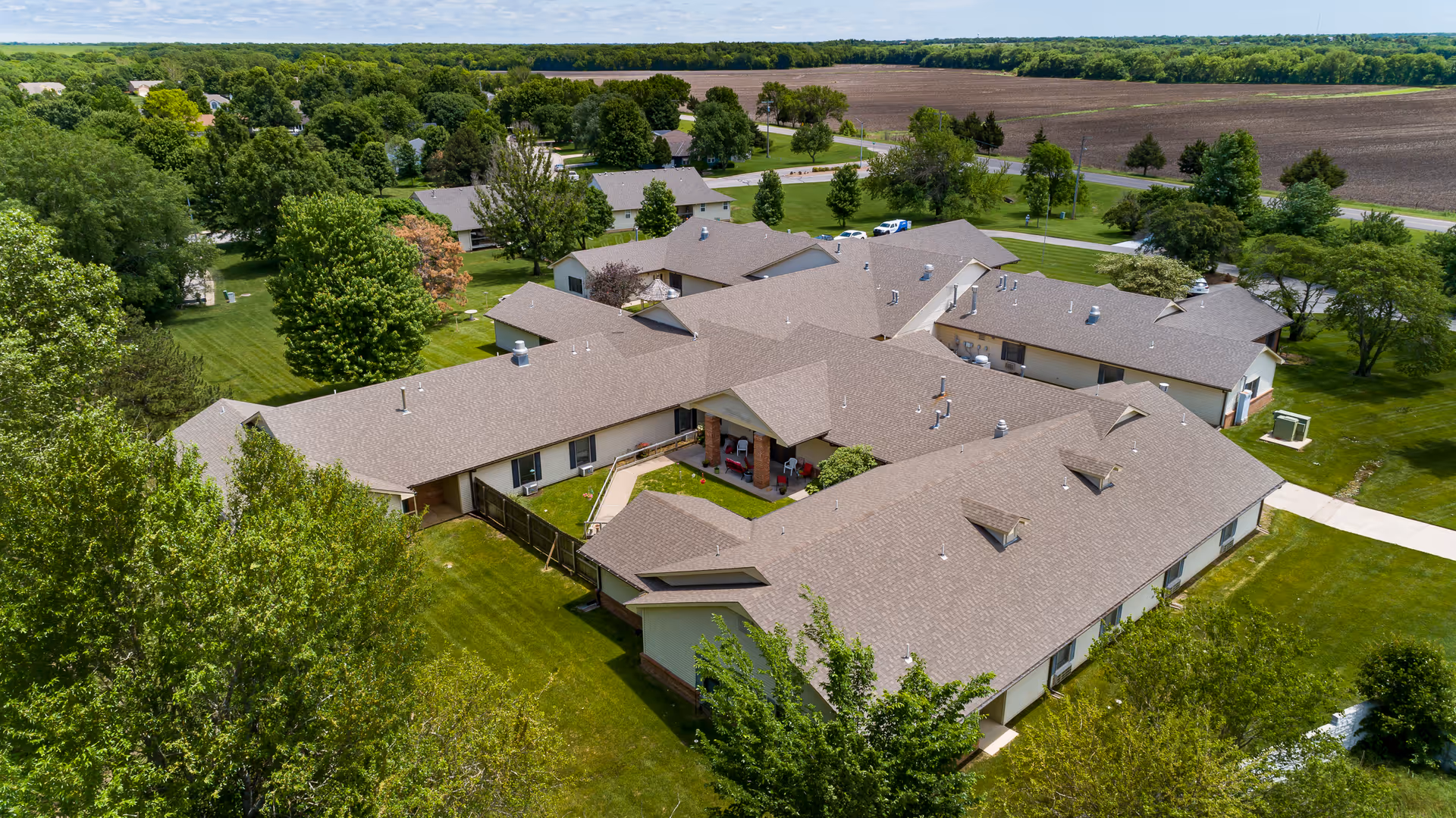 Aerial view of Homestead Senior Living of El Dorado showing a large, single-story building complex surrounded by green lawns, trees, and open fields under a partly cloudy sky.