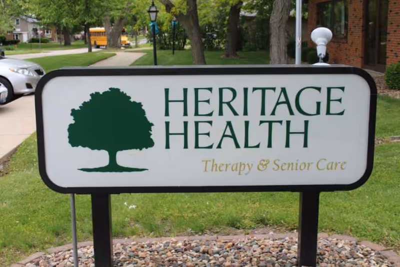Outdoor sign for Heritage Health Therapy & Senior Care with a green tree logo, situated on a grassy area with a sidewalk, parked cars, and a school bus in the background.