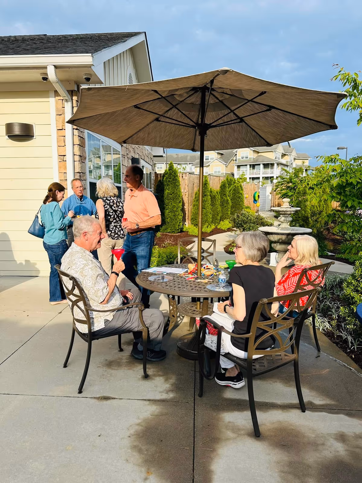 A group of elderly people socializing outdoors at a senior living facility. Three people are seated around a round metal table with an umbrella, holding colorful drinks, while four others stand nearby engaged in conversation. The setting includes a paved patio area, greenery, and a water fountain in the background.