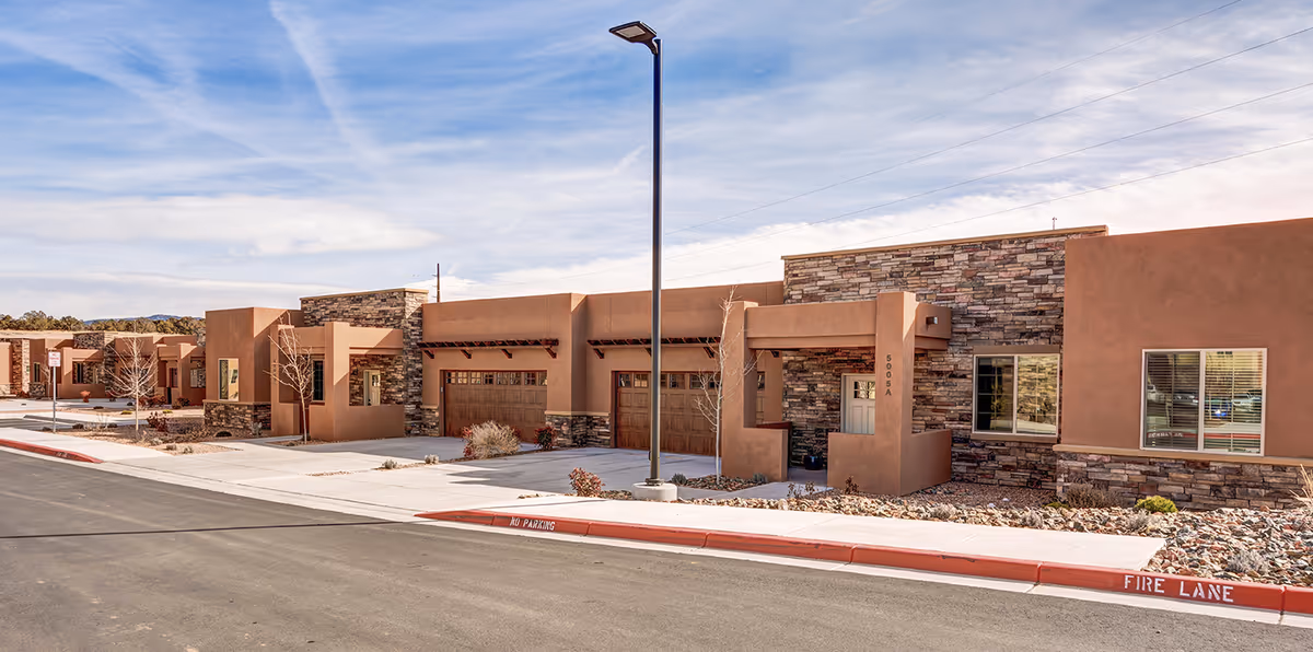 Exterior view of a modern senior living facility with southwestern-style architecture featuring tan stucco walls, stone accents, and wooden garage doors. The building is single-story with a clean sidewalk and a street marked with 'No Parking' and 'Fire Lane'. A street lamp stands in front of the building under a partly cloudy sky.