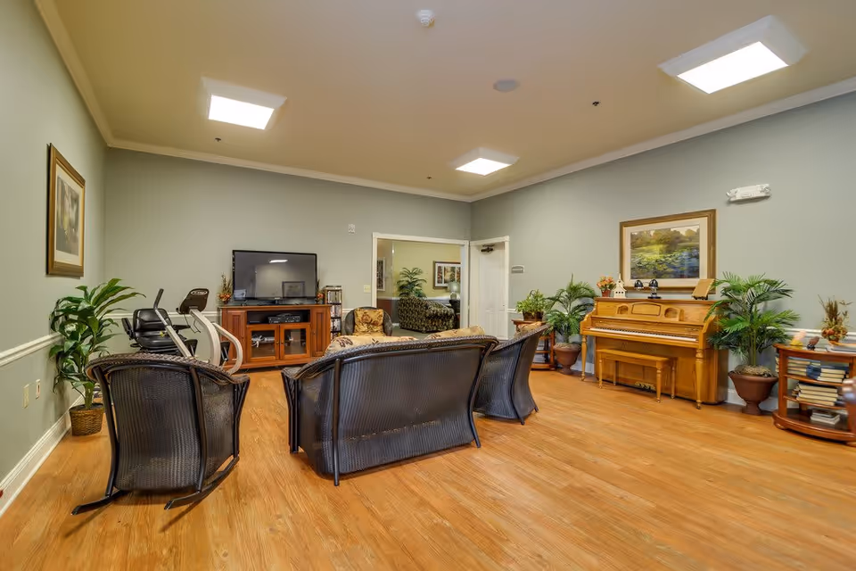 A cozy senior living common area with wooden flooring, a piano against the wall, a flat-screen TV on a wooden cabinet, exercise equipment, several wicker chairs and a sofa, potted plants, and framed artwork on the walls.