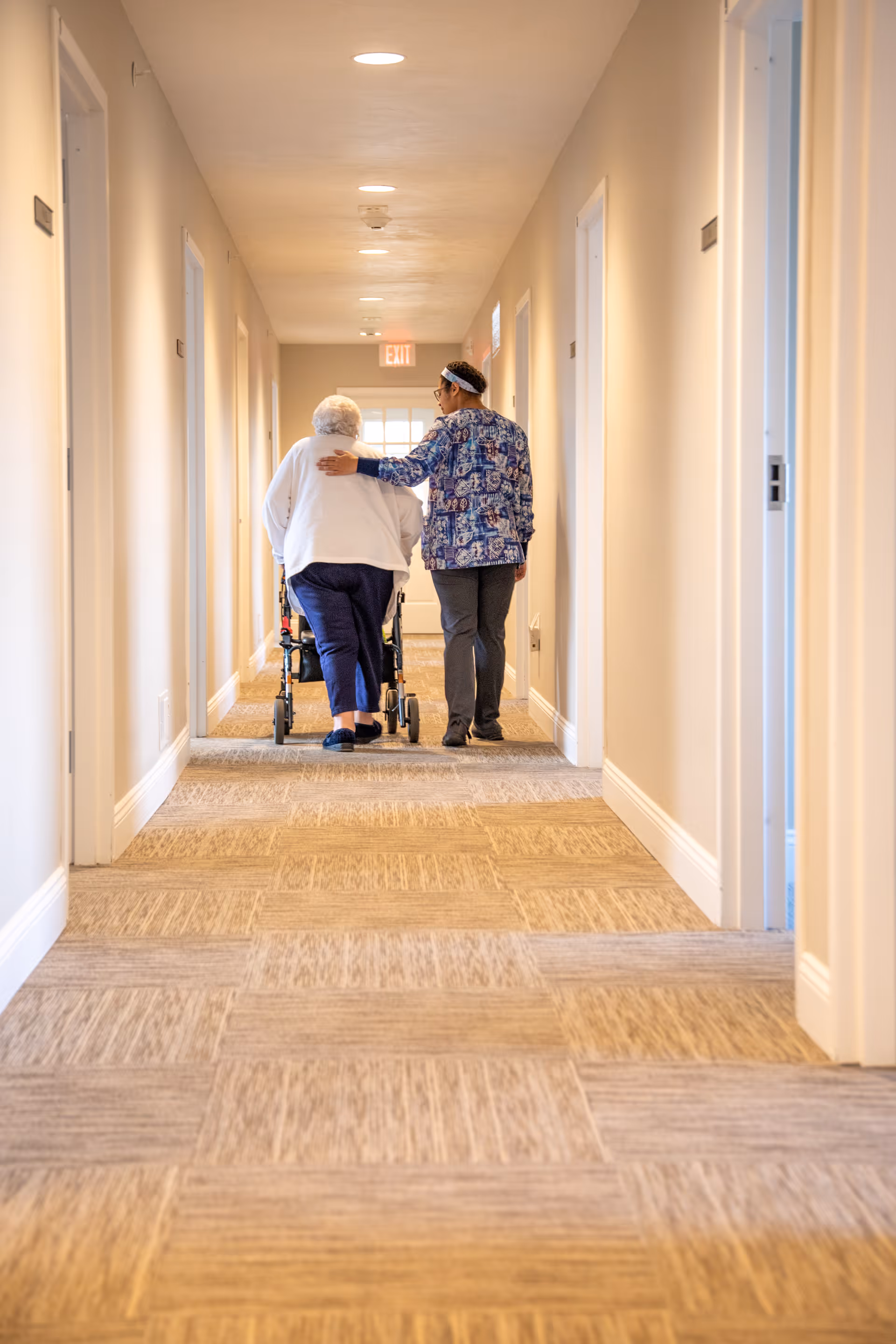 A caregiver walking alongside an elderly person using a walker down a well-lit hallway in a senior care facility. The caregiver has their arm gently around the elderly person's shoulders, providing support and companionship.