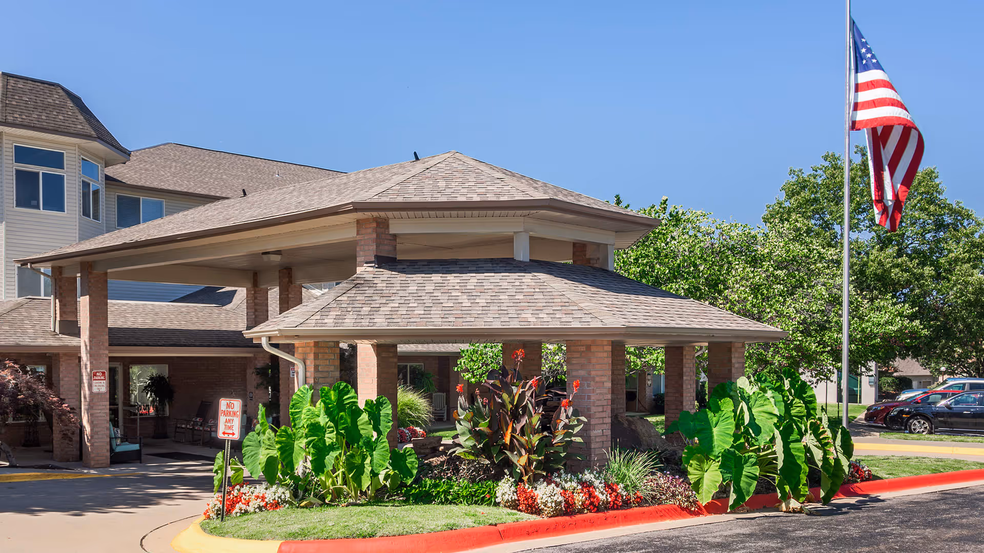 Exterior view of the entrance to The Gardens at Arkanshire Senior Living facility, featuring a covered drop-off area with brick pillars, lush green plants, colorful flowers, an American flag on a flagpole, and a clear blue sky.