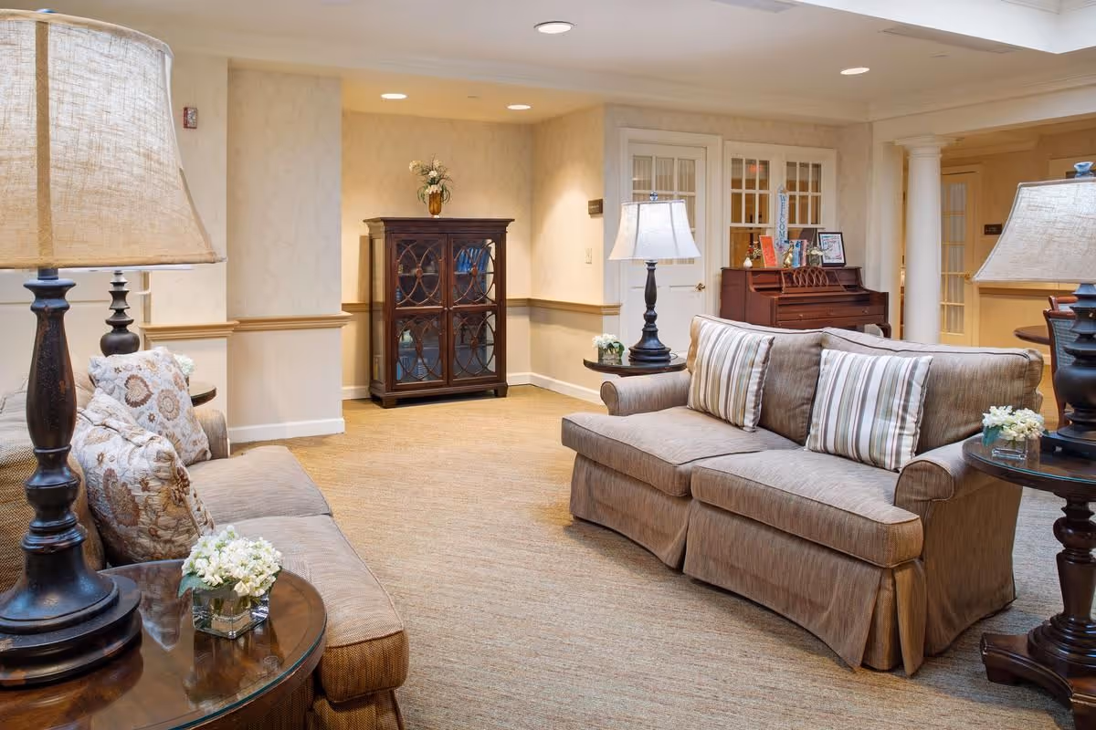 A cozy living room area with two beige sofas adorned with patterned and striped pillows. There are two dark wooden side tables with lamps and small flower arrangements. In the background, there is a wooden cabinet with glass doors and a piano with framed pictures and a welcome sign on top. The room has beige walls, carpeted flooring, and recessed ceiling lights.