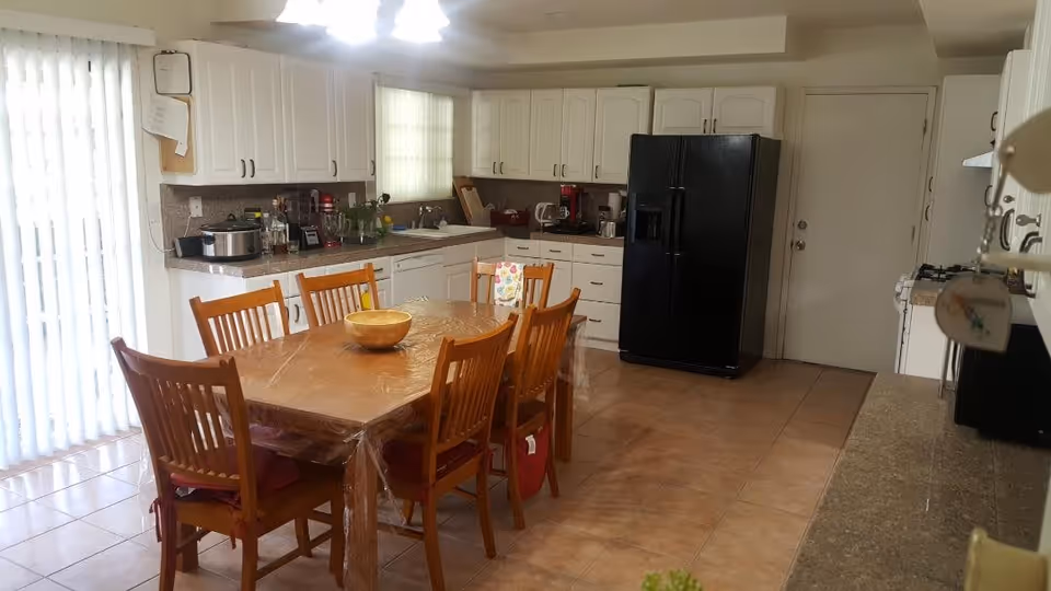 A spacious kitchen with white cabinets, a black refrigerator, a stove, and a dishwasher. There is a wooden dining table covered with a clear plastic tablecloth and six wooden chairs around it. The floor is tiled, and there is a sliding glass door with vertical blinds on the left side letting in natural light.