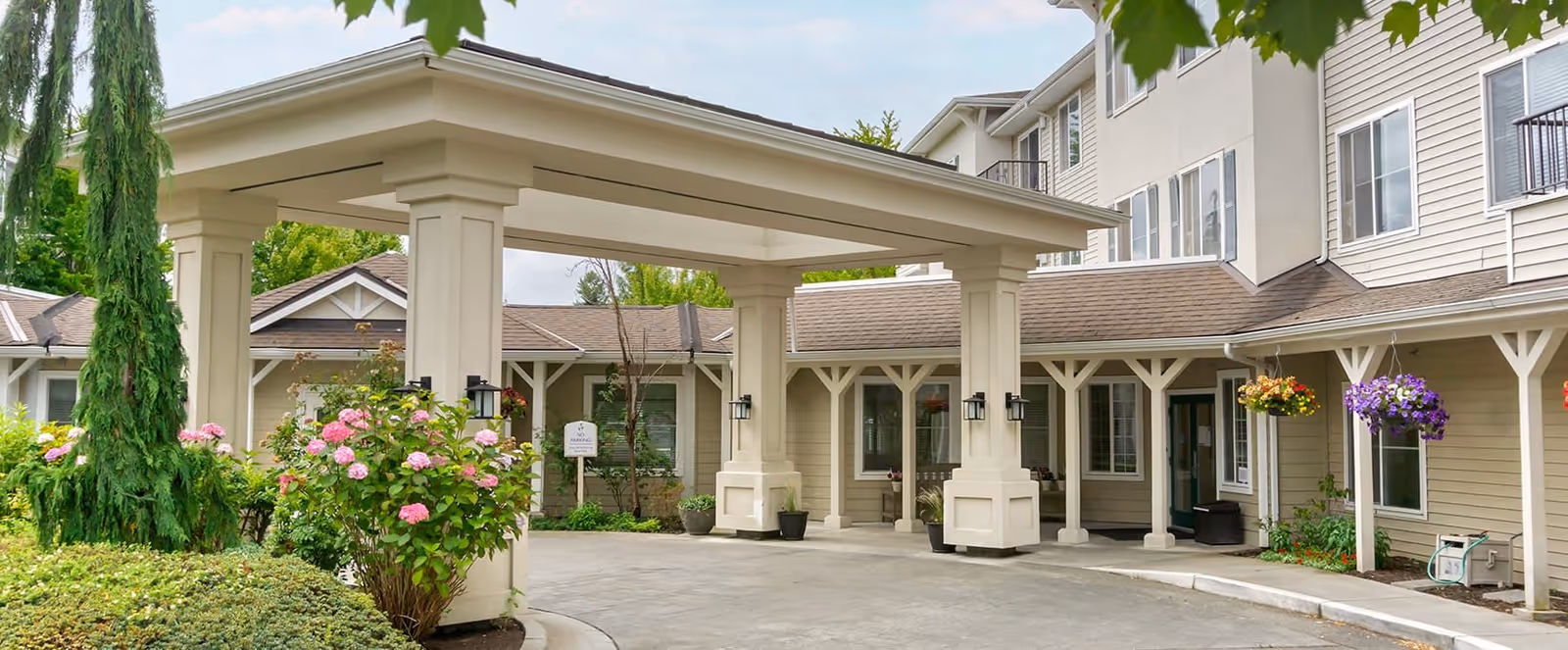 Entrance area of a senior living facility with a covered drop-off zone supported by large columns. The building exterior is beige with white trim, and there are hanging flower baskets and well-maintained landscaping including bushes and trees.