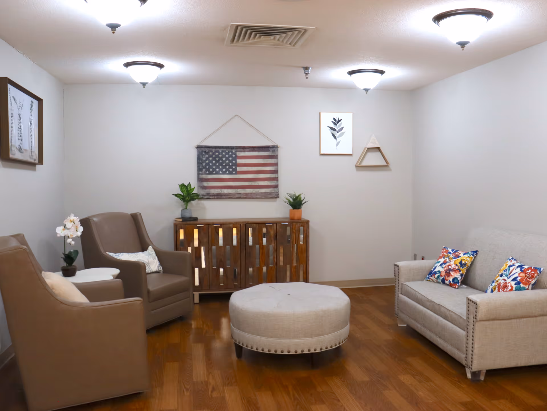 A cozy living room with two brown armchairs on the left, a beige sofa with colorful floral pillows on the right, and a round beige ottoman in the center. A wooden cabinet with plants and an American flag hanging above it is against the back wall. The walls are light-colored with framed artwork, and the floor is wooden.