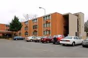 Exterior view of a three-story brick apartment building with several parked cars in front under a cloudy sky. The building has multiple windows and an external stairwell on the right side.
