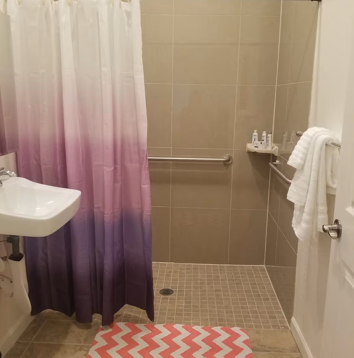 A bathroom with a walk-in shower featuring beige tiles and a metal grab bar. There is a white sink on the left side and a shower curtain with a gradient from white to purple. A pink and white chevron-patterned bath mat is on the floor. White towels hang on a metal bar attached to the door, and small toiletries are placed on a corner shelf inside the shower.