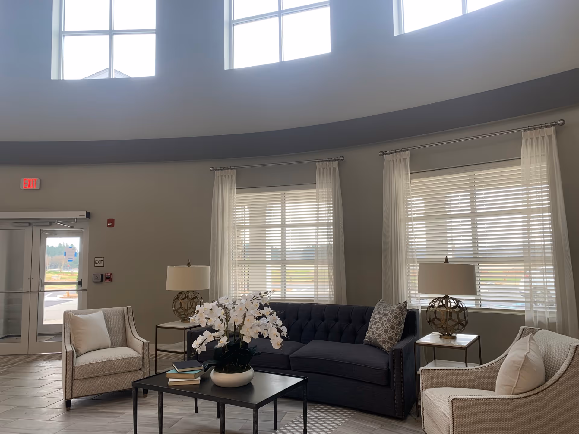 A bright and spacious living room area with a dark blue sofa, two beige armchairs with white cushions, a black coffee table with a white orchid plant and some books, two side tables with decorative lamps, large windows with white blinds and sheer curtains, and a glass door exit with an exit sign above it.