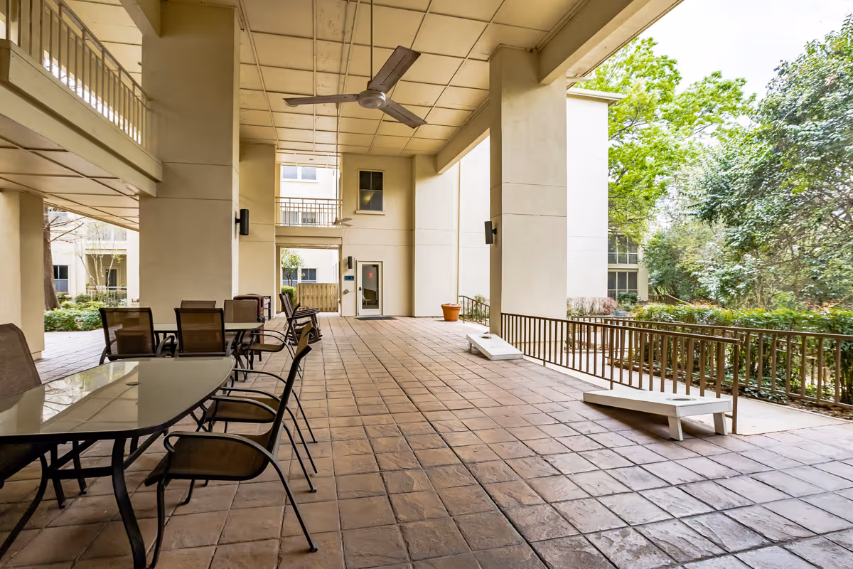 Covered outdoor patio area with tiled floor, ceiling fans, glass tables with chairs, and two cornhole game boards. The space is adjacent to a garden with trees and bushes, and there are railings along the edge of the patio.