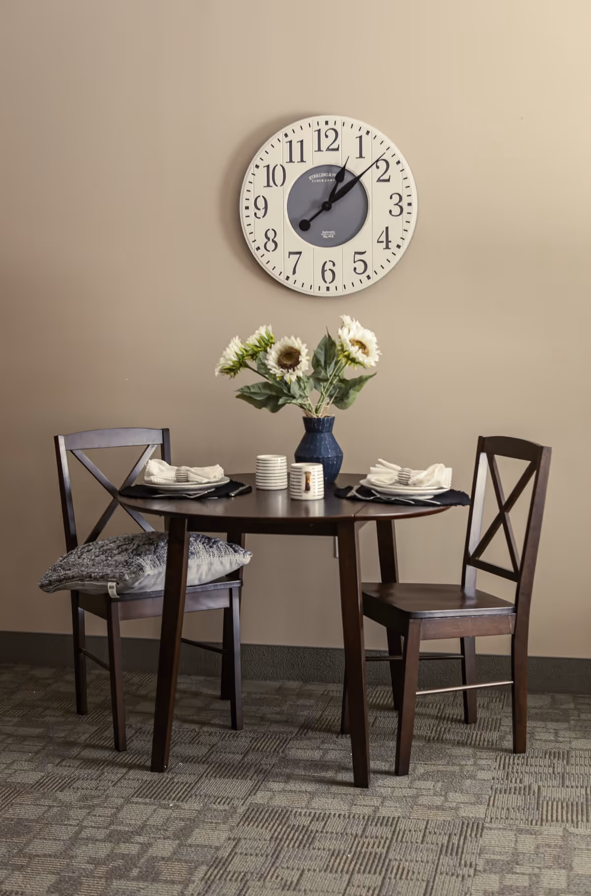 A small dining area with a round dark wooden table set for two, featuring plates, napkins, and utensils. A blue vase with white sunflowers is placed in the center of the table. Two wooden chairs with cushions are positioned on either side of the table. A large round wall clock with black numbers and hands hangs on the beige wall behind the table.