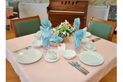 A dining table set for four with plates, cups, folded blue napkins and a floral centerpiece, with green upholstered chairs and a piano in the background.