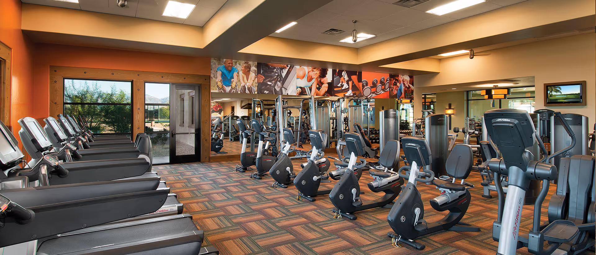 Interior view of a fitness center with multiple treadmills on the left and several stationary exercise bikes and other workout equipment on the right. The room has large windows and glass doors letting in natural light, with colorful carpet flooring and a ceiling with recessed lighting. There are motivational fitness posters above the mirrors on the far wall.