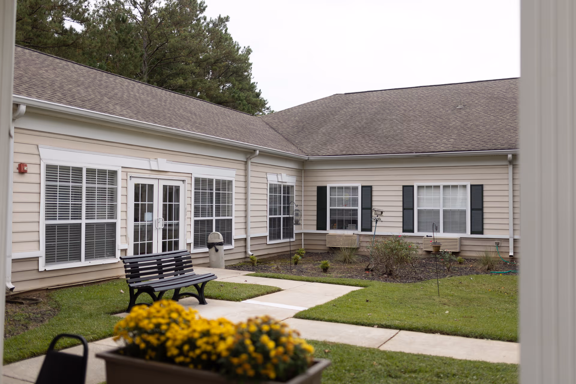 Outdoor courtyard area of a senior living facility with beige siding buildings, multiple windows with white blinds, a black bench on a concrete pathway, green grass, and yellow flowers in a planter in the foreground.