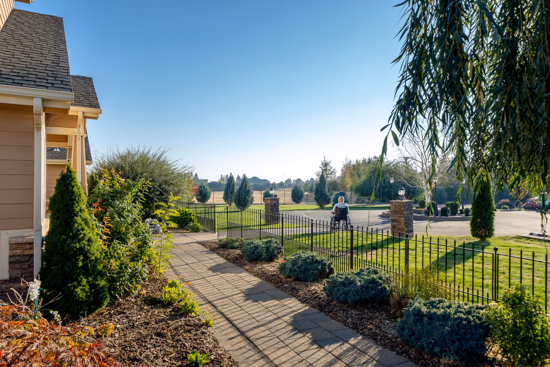 A sunny outdoor scene at Pleasant Comfort AFH showing a paved walkway alongside a building with shrubs and small trees. A person in a wheelchair is seen in the distance near a black metal fence and stone pillars, with a parking area and more trees in the background under a clear blue sky.