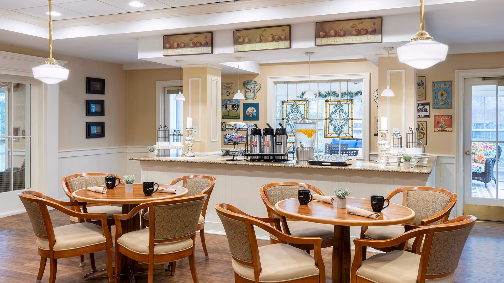 A cozy dining area with two round wooden tables, each surrounded by four cushioned chairs. The tables are set with black mugs, napkins, and small potted plants. In the background, there is a counter with coffee dispensers, a water dispenser with lemon slices, and decorative items. The room has warm beige walls, framed artwork, and pendant lights hanging from the ceiling. A door and windows with stained glass panels are visible, leading to an outdoor seating area.