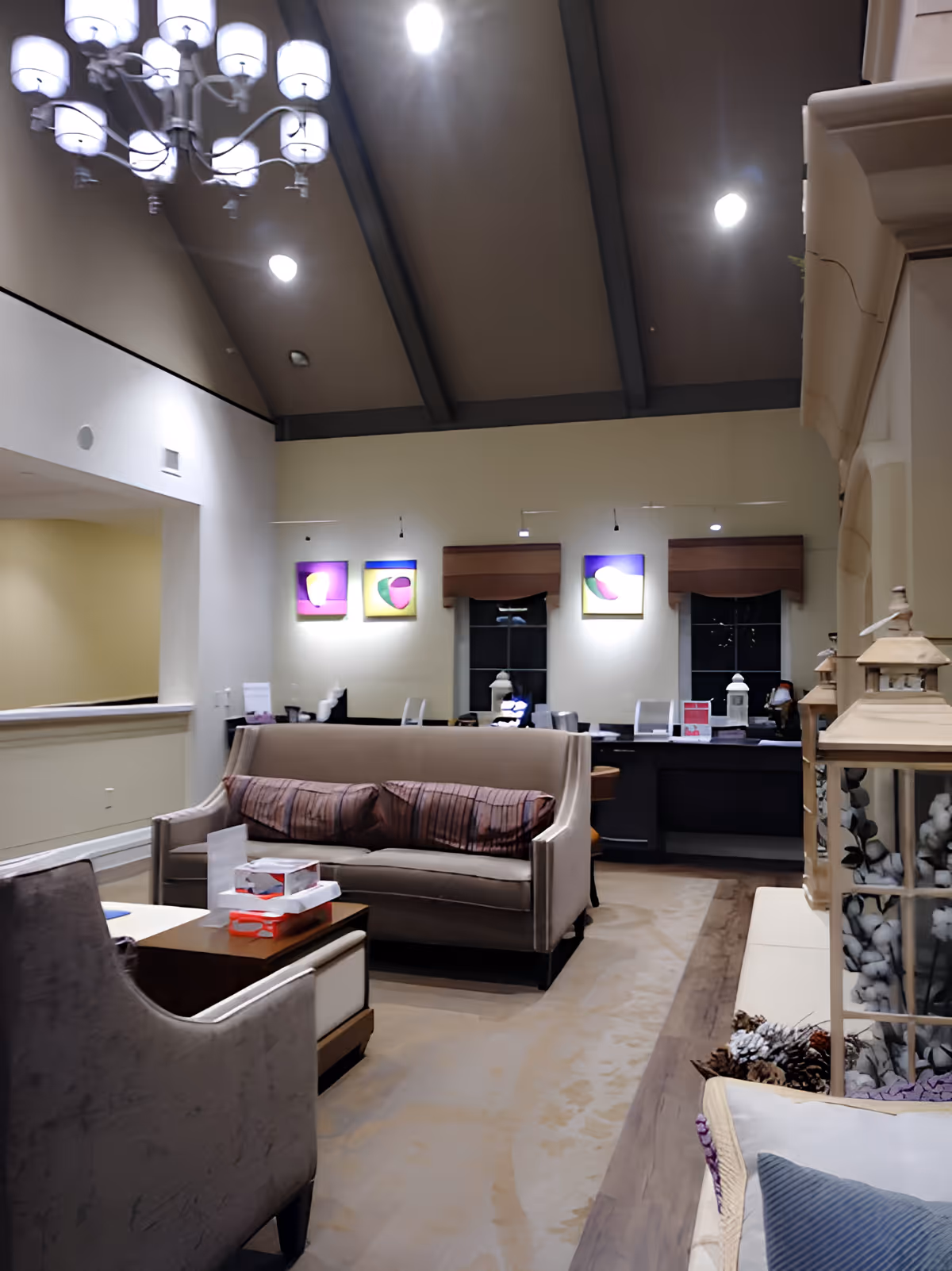 Interior view of a living room area in a senior living facility with a beige sofa, armchair, coffee table with tissue boxes, decorative lanterns, and colorful artwork on the wall under a high ceiling with exposed beams and a chandelier.
