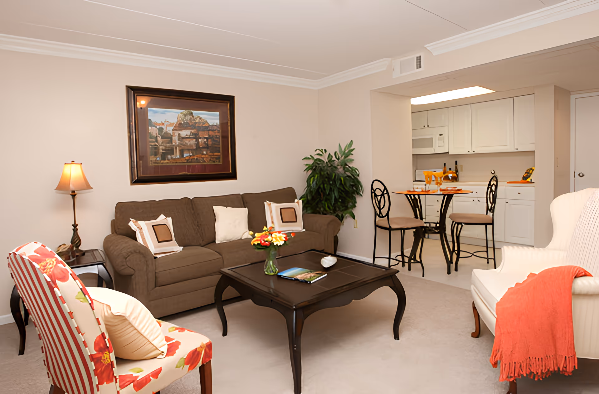 A cozy living room with a brown sofa adorned with decorative pillows, a floral patterned chair, and a white armchair with a red throw blanket. A dark wooden coffee table with a vase of flowers and a book sits in the center. The room has beige walls with a framed painting, a floor lamp, and a green potted plant. In the background, there is a small dining area with a round table and two chairs, adjacent to a white kitchen with cabinets and appliances.