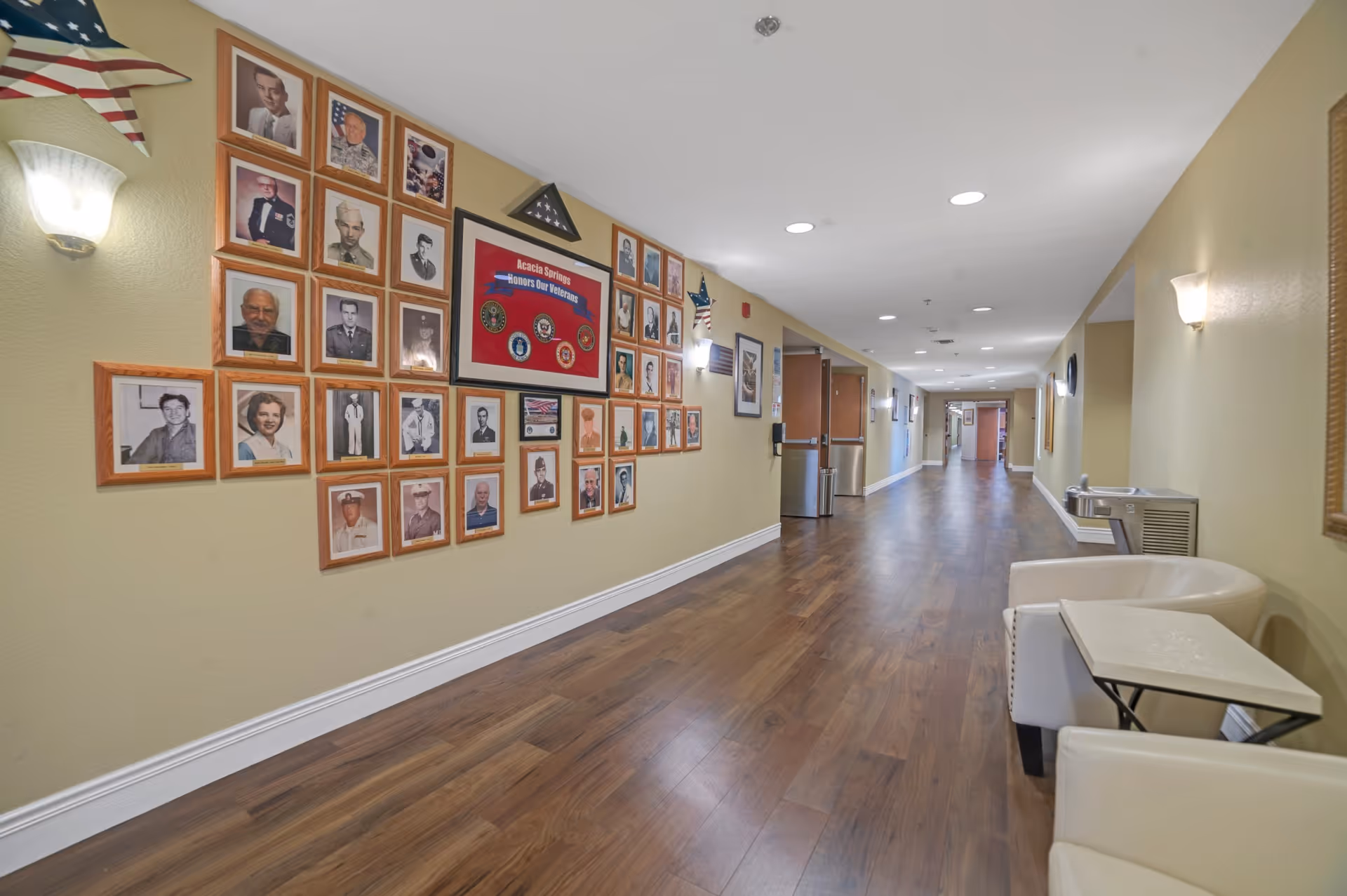 A long hallway in a senior living facility with wooden flooring and beige walls. On the left wall, there is a display of framed photographs of veterans along with a framed flag and a sign that reads 'Acacia Springs Honors Our Veterans.' There are wall-mounted lights and an American flag above the display. On the right side, there are two white chairs with a small table between them and a water fountain further down the hallway.