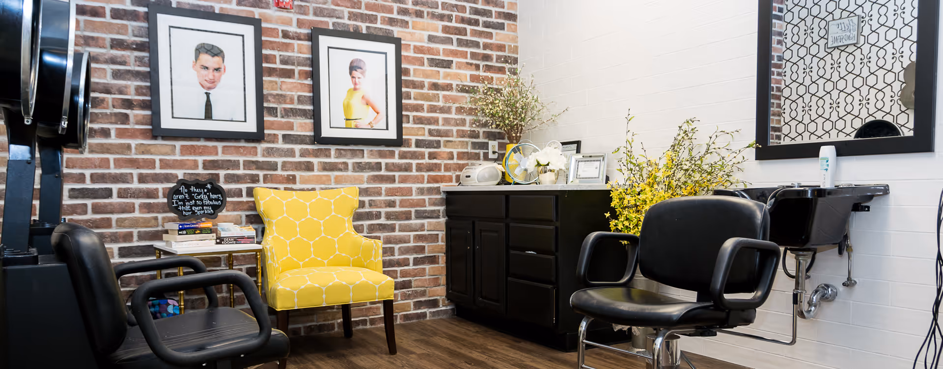 Small salon-style interior with brick accent wall, salon chairs, a yellow patterned armchair, and a black vanity with a mirror and framed artwork.