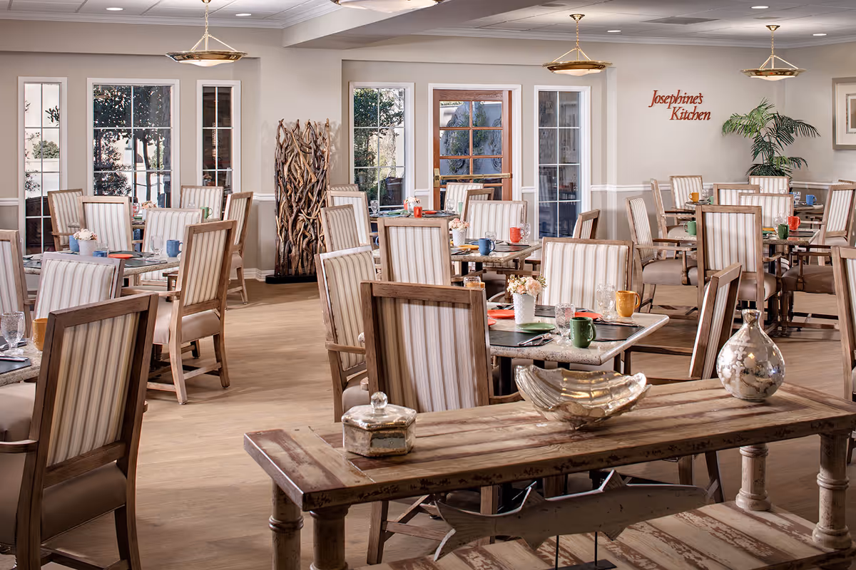A bright and spacious dining room with multiple tables and wooden chairs featuring striped upholstery. Each table is set with colorful mugs, plates, glasses, and small flower arrangements. The room has large windows and a glass door letting in natural light. On the wall, there is a sign that reads 'Josephine's Kitchen'. Decorative items including a wooden sculpture and metallic vases are placed on a rustic wooden table in the foreground.