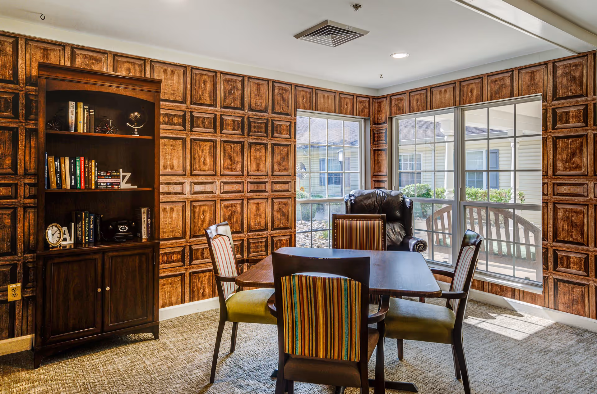 A cozy interior room with wood-paneled walls and a large window overlooking an outdoor area. The room features a dark wooden table surrounded by four chairs with striped upholstery, a leather armchair near the window, and a wooden bookshelf filled with books and decorative items.