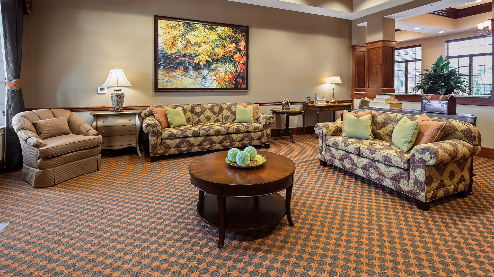 A cozy living room with two patterned sofas adorned with green and orange pillows, a cushioned armchair, a round wooden coffee table with decorative items, side tables with lamps, a large window with blinds, and a colorful painting of autumn trees on the wall.