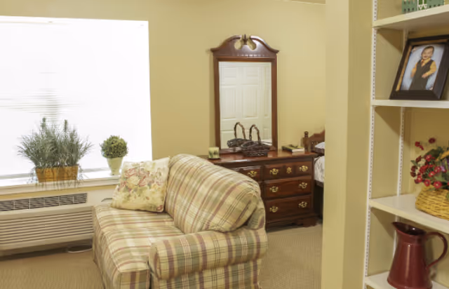 A cozy room with a plaid upholstered loveseat, a floral pillow, a wooden dresser with a mirror, and a window with potted plants on the windowsill. A bookshelf with framed photos and decorative items is partially visible on the right.