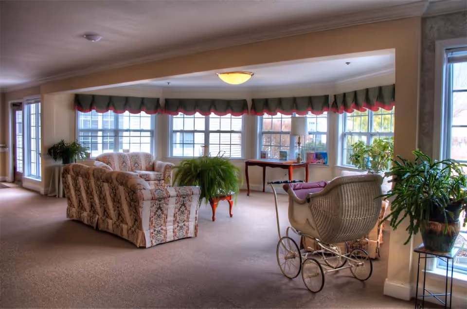 A cozy living room area with floral patterned sofas, a vintage baby carriage, several potted plants, and large windows with green and red valances allowing natural light to fill the space. There is also a wooden table with a lamp and decorative items near the windows.