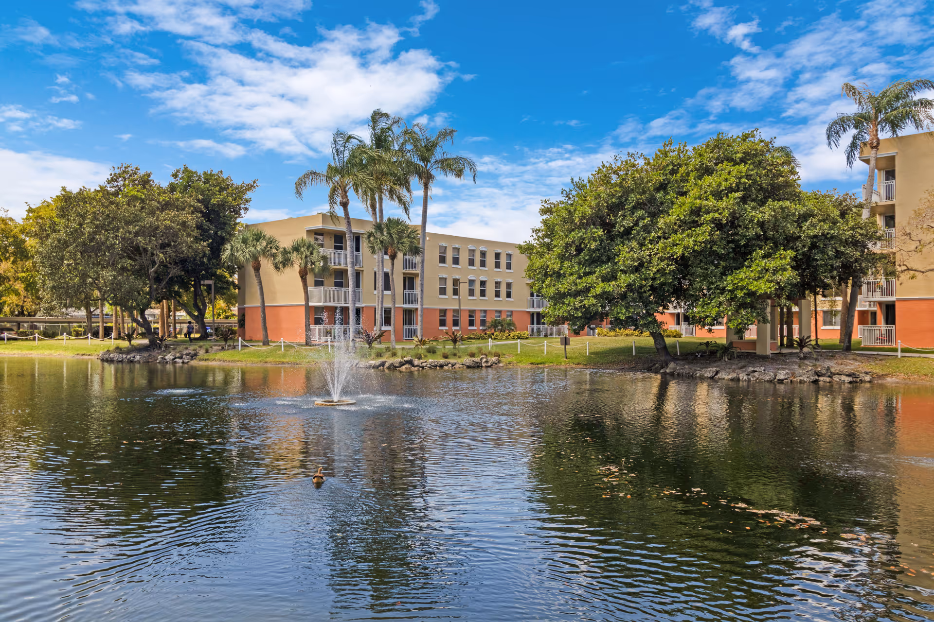A serene outdoor scene at Five Star Premier Residences of Hollywood featuring a pond with a water fountain in the center, surrounded by lush green trees and palm trees. In the background, there is a multi-story residential building with beige and orange exterior walls under a partly cloudy blue sky.