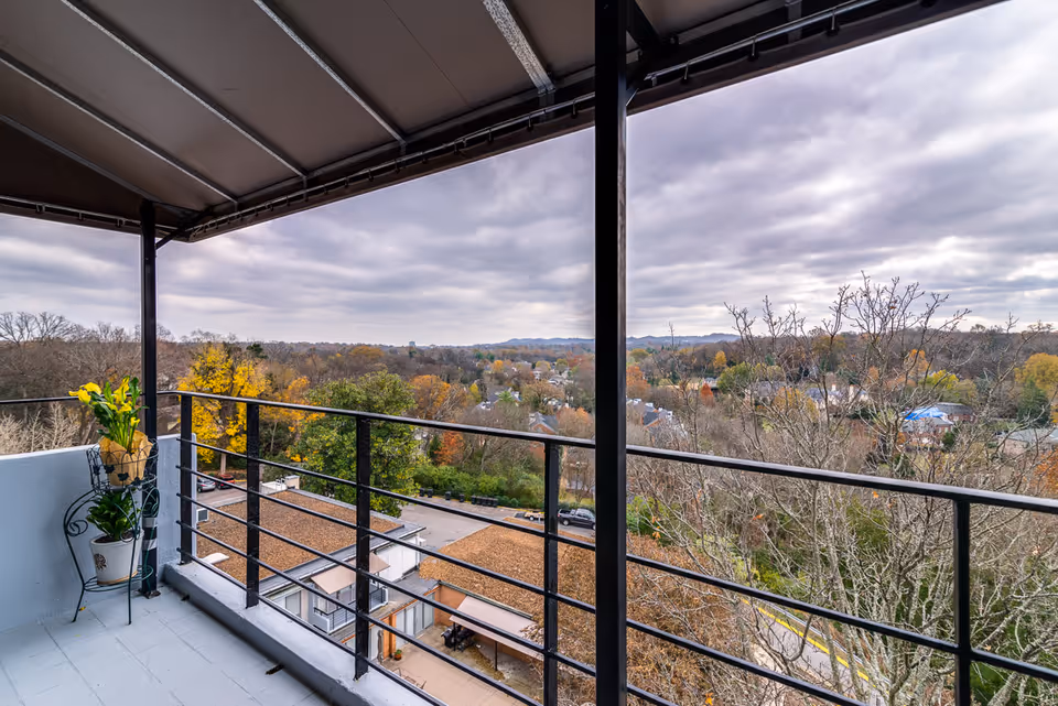 View from a covered balcony overlooking a residential area with trees showing autumn colors under a cloudy sky. The balcony has a black metal railing and two potted plants on a decorative stand.