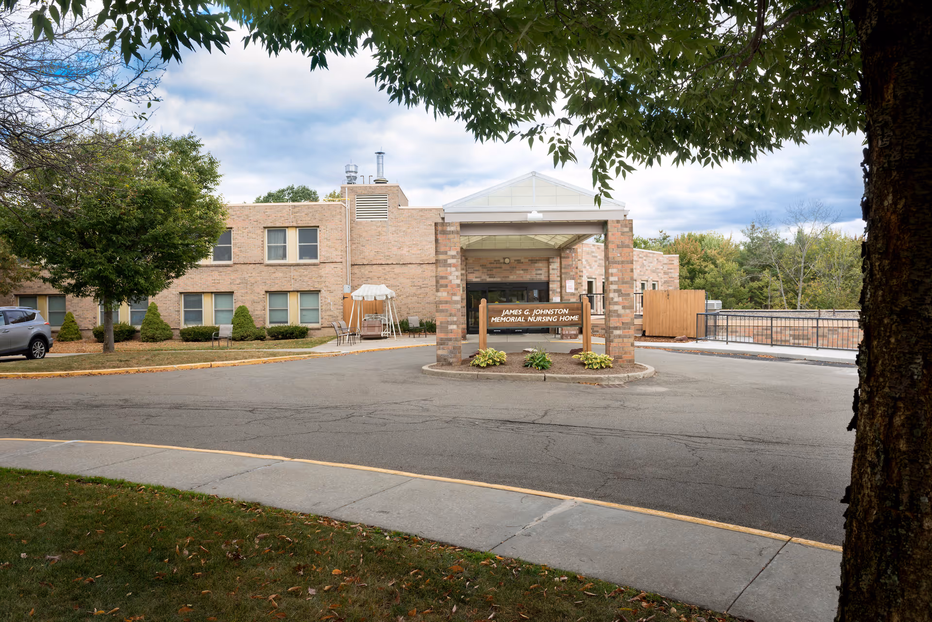 Exterior view of the James G. Johnston Memorial Nursing Home building at United Methodist Homes Hilltop Campus, showing a brick structure with a covered entrance, a driveway, some trees, and a parked car.
