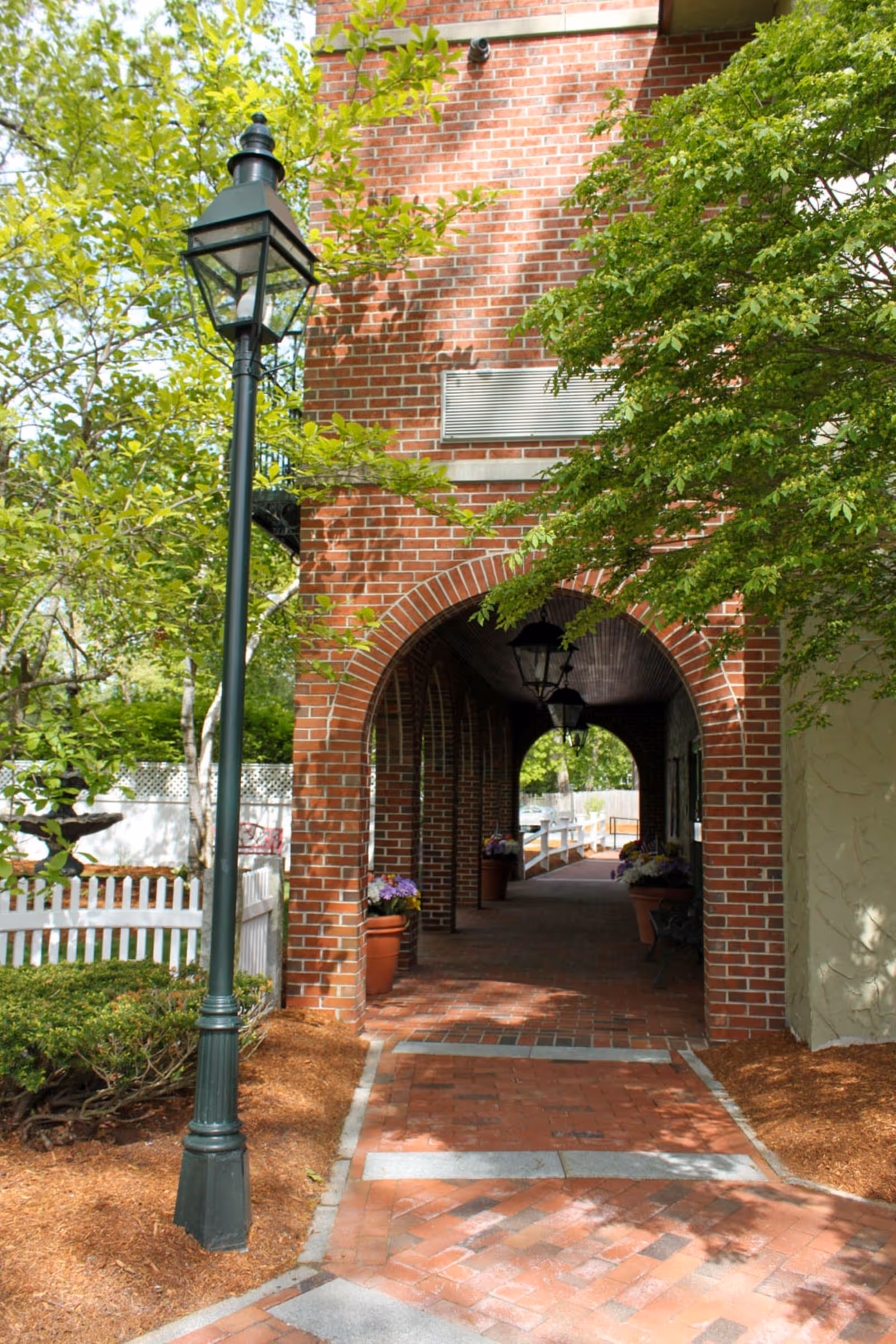 A brick archway walkway with hanging lanterns, potted plants, and a black street lamp on the left side. The area is surrounded by green trees and bushes, with a white picket fence visible in the background.