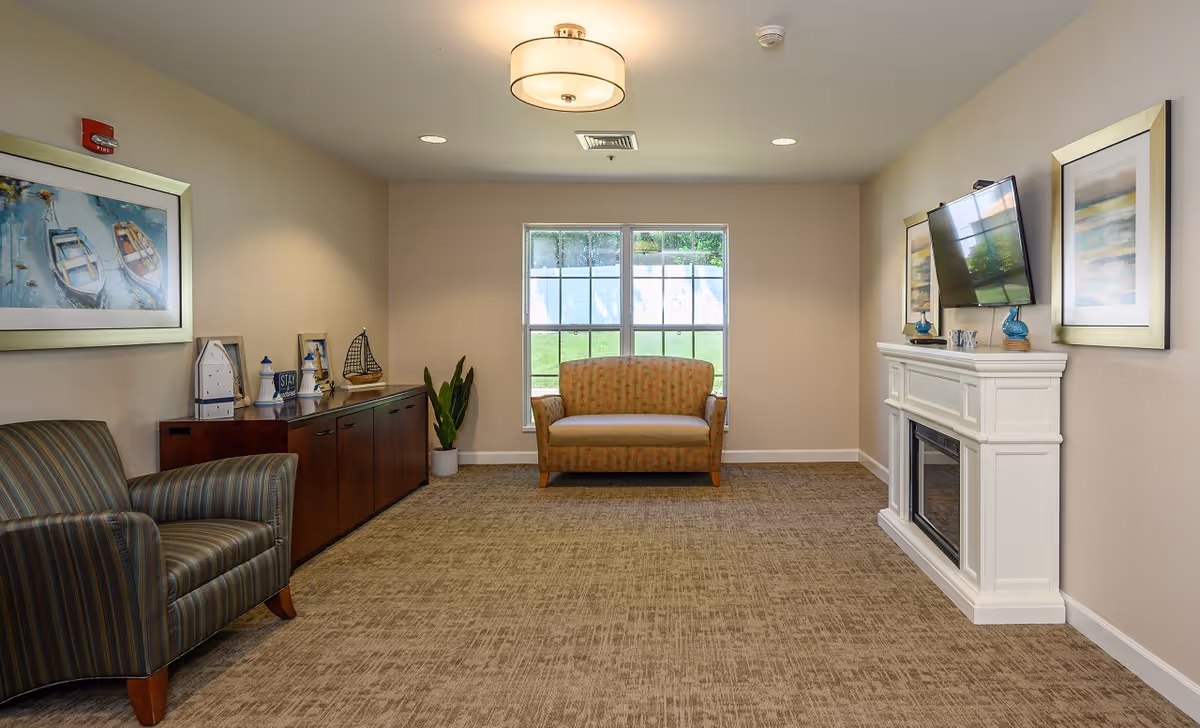 A cozy living room with a striped armchair on the left, a small loveseat in front of a window, and a white fireplace with a mounted flat-screen TV on the right. The room has beige walls, carpeted floor, and decorative framed artwork on the walls. A wooden cabinet with nautical-themed decor is along the left wall.