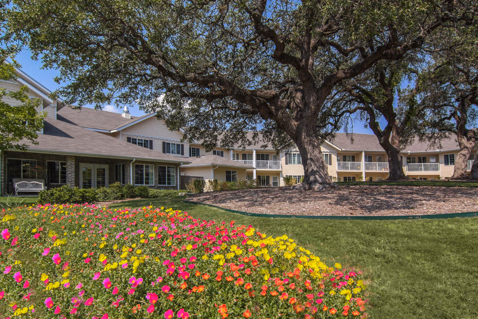 A senior living facility building with beige siding and white trim, surrounded by large mature trees and a well-maintained garden with colorful flowers in the foreground under a clear blue sky.