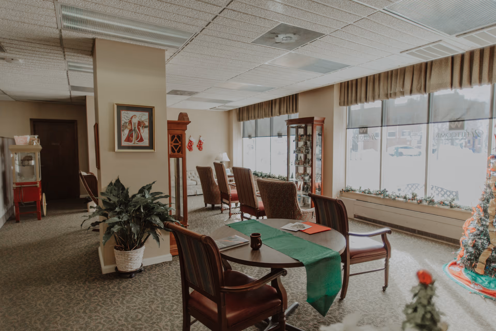 A cozy senior living common area with a round table covered by a green runner and surrounded by four chairs. The room has large windows letting in natural light, a decorated Christmas tree, a grandfather clock, a display cabinet, and comfortable armchairs arranged near the windows. There is a potted plant and a popcorn machine in the background, with holiday stockings hanging on the wall.