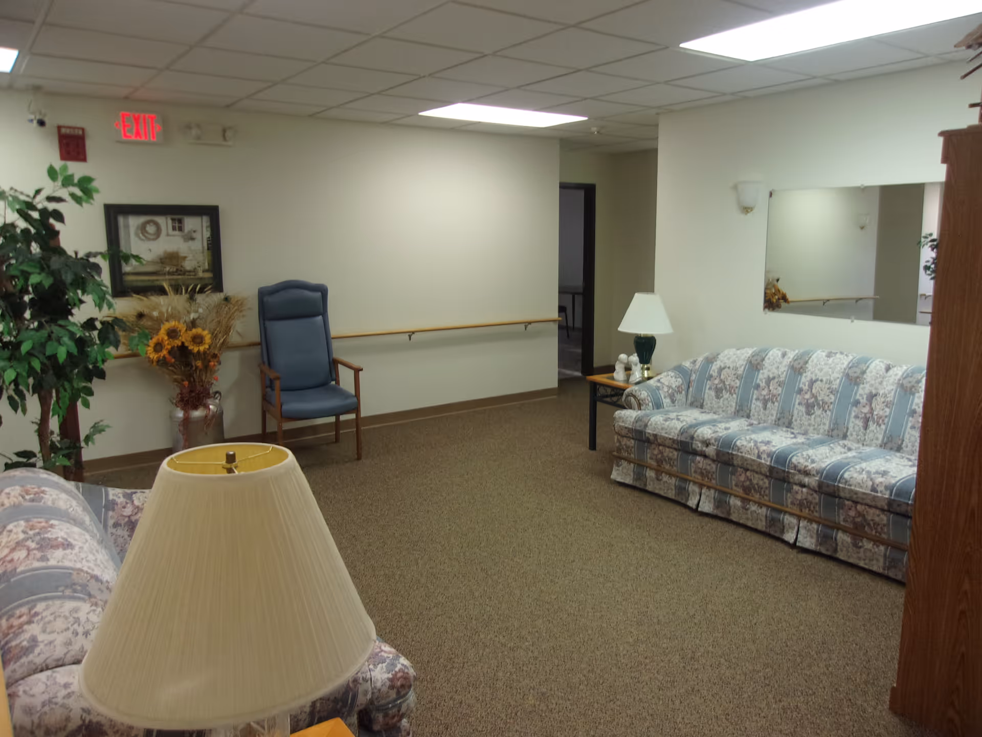 Carpeted common sitting room with floral-pattern sofas, chairs, lamps, plants and an EXIT sign.