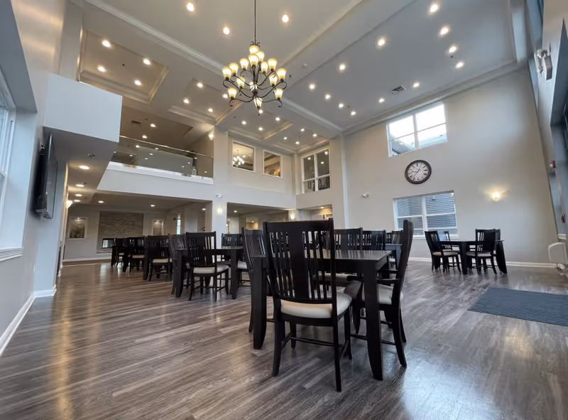 A spacious dining room with multiple dark wooden tables and chairs arranged neatly on a wood floor. The room has high ceilings with recessed lighting and a large chandelier. There are windows on the walls allowing natural light in, and a large clock is mounted on one wall. The room has a modern and clean design with light-colored walls.
