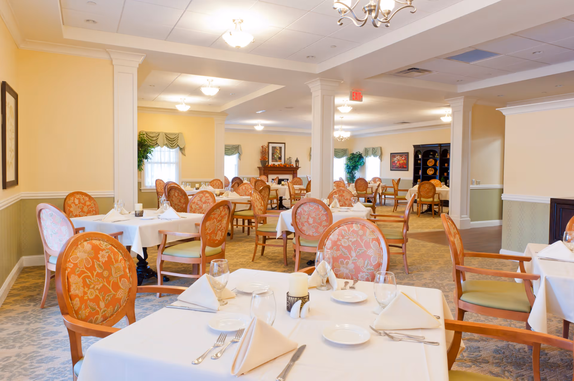 A bright and elegant dining room with multiple tables covered in white tablecloths, set with plates, glasses, silverware, and folded napkins. The chairs have wooden frames with floral upholstery. The room features soft yellow walls, white trim, decorative columns, and several light fixtures on the ceiling. There are windows with green valances and framed artwork on the walls.