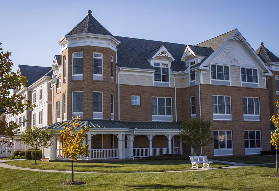 Exterior view of a large, multi-story senior living facility building with brick and white siding, featuring a turret, multiple windows, a covered porch, and a well-maintained lawn with small trees and a white bench.